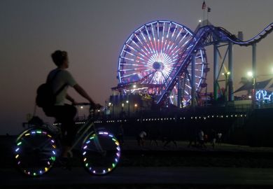 A cyclist rides along a path with lights on the wheels on Santa Monica Pier with Ferris wheel in multiple lights to illustrate NIH looks to slim down peer review in bid for equity
