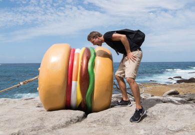 A person  mocks taking a bite out of "What a Tasty Looking Burger" by James Dive at Sculpture By The Sea in Sydney, Australia to illustrate Campus catering ‘too expensive and unhealthy’