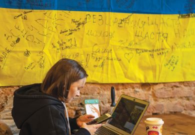 A woman works on a laptop in a room with a  Ukrainian flag on the wall to illustrate International help ‘must not kill Ukrainian system with kindness’