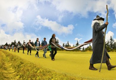 A man dressed as a grandfather leads people carrying a stick with the people holding a long knitted scarf
