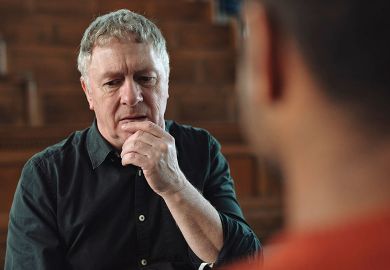 Man with grey hair and black shirt holding hand at mouth whilst thinking and talking to male