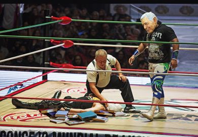 In a wrestling ring filled with books, one man lies prone and is being counted out while his masked opponent prowls In a wrestling ring filled with books, one man lies prone and is being counted out while his masked opponent prowls
