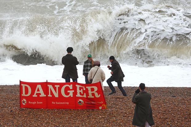 People walk on the beach at high tide, during strong winds in Brighton, United Kingdom, with a sign warning of dangerous seas. To illustrate English universities being ‘unprepared’ as new free speech rules loom. People walk on the beach at high tide, during strong winds in Brighton, United Kingdom, with a sign warning of dangerous seas. To illustrate English universities being ‘unprepared’ as new free speech rules loom.