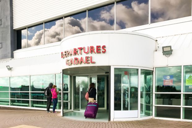 Cardiff, Wales - June 2019:: Person pulling a suitcase entering the terminal building at Cardiff Wales Airport. Blue sky and clouds are reflected in the windows. The airport is publicly owned by the Welsh Government.