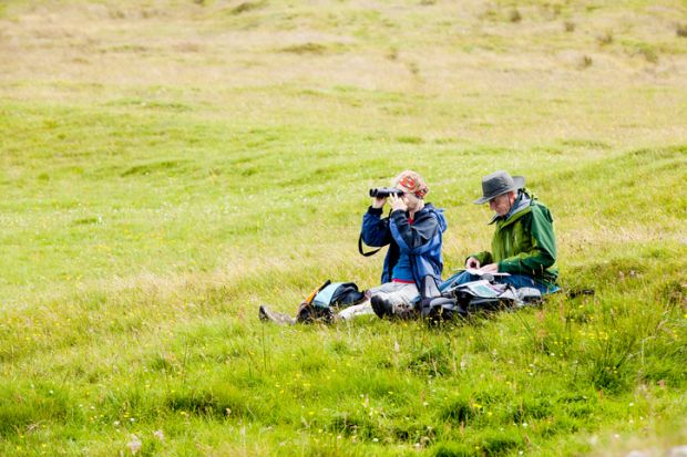 Two senior adults, a man and a woman, sit on the ground in a green meadow in Skye, Scotland Two senior adults, a man and a woman, sit on the ground in a green meadow in Skye, Scotland