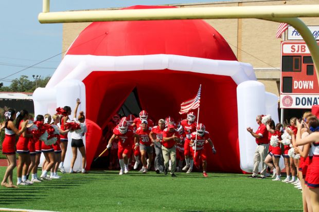 High school football team entering the field through a blow up football helmet tunnel with cheerleaders in a line cheering.