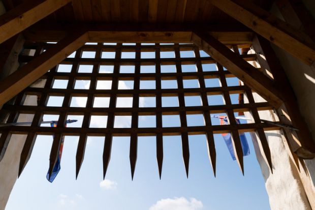 View of a medieval wooden portcullis gate with blue sky and flags in the background