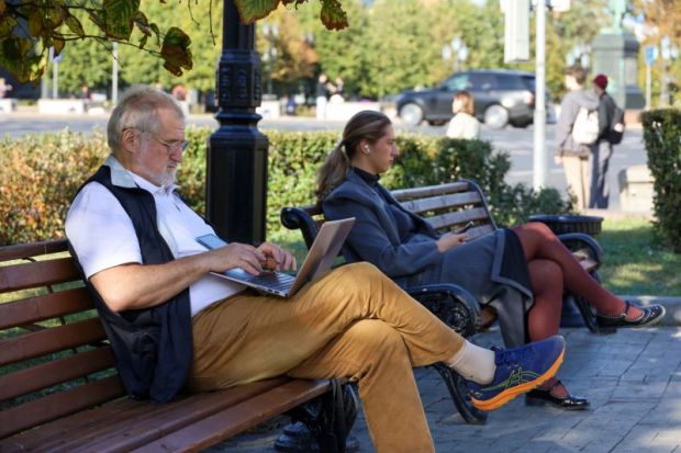Man sitting with laptop on wooden bench on a street.