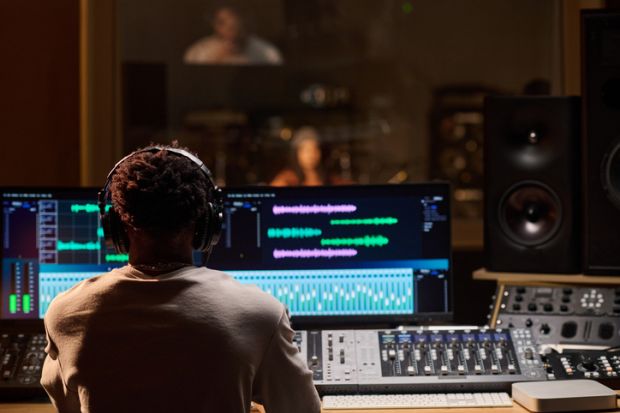 Man wearing headphones operating audio mixing console and computer monitors in professional recording studio, observing musician performing through glass window in background