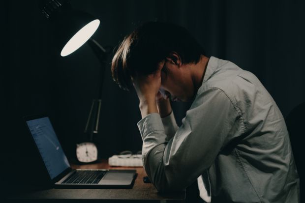 A man working on a laptop holds his head late at night, illustrating overwork
