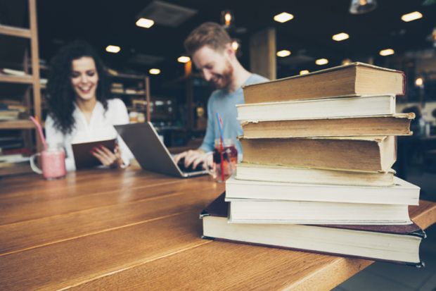 Students sitting at a table alongside a pile of books Students sitting at a table alongside a pile of books