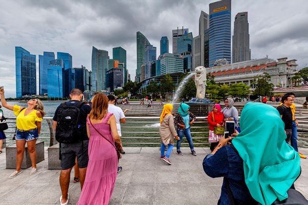Tourists of various nationalities pose for photos in front of the Merlion statue and Singapore skyline, illustrating Singapore’s international nature