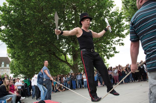Street performer thrills the crowd with his knife act whilst balancing on a rope slack line being held up by members of the audience to illustrate UKRI ‘must regain trust’ of equality committee in Donelan row