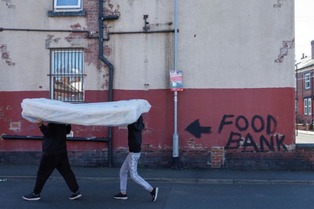 Two men carry a mattress over their heads past a terraced house, the walls of which bare graffiti reading "food bank" to illustrate Cost of living crisis bites for PhDs on ‘inadequate’ stipends