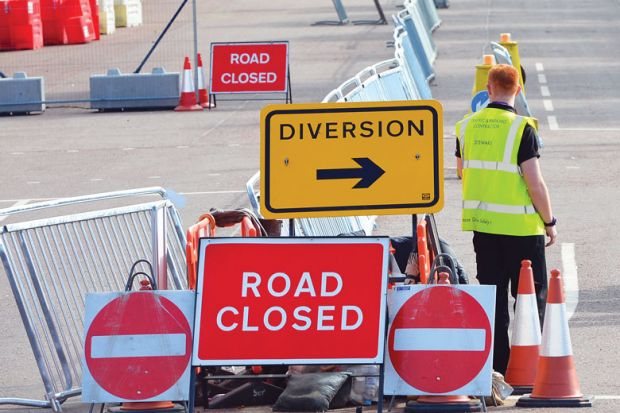 Worker standing with road-signs as a metaphor for shortages at labs caused by border snarls and new rules.